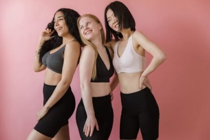 Three women in athletic wear pose joyfully against a pink background, celebrating body positivity.